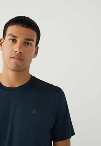 Marineblå sports T-shirt med sorte skulderdetaljer og et lille logo på brystet. Glat tekstur med korte ærmer og rund hals.