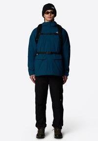 Teal waterproof jacket with black belt, two front pockets, paired with black pants and sturdy boots. Accessories include black beanie and sunglasses.