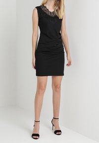 Robe noire sans manches avec un col en dentelle, une coupe ajustée et un ourlet longueur genou. Texture lisse avec une silhouette élégante.