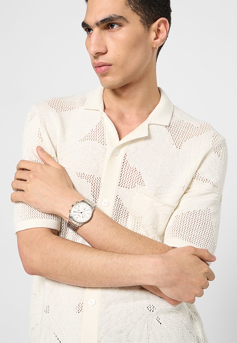 Chemise blanche en tricot à manches courtes avec un motif géométrique et une poche à l'avant, assortie à une montre-bracelet en argent avec un grand cadran.