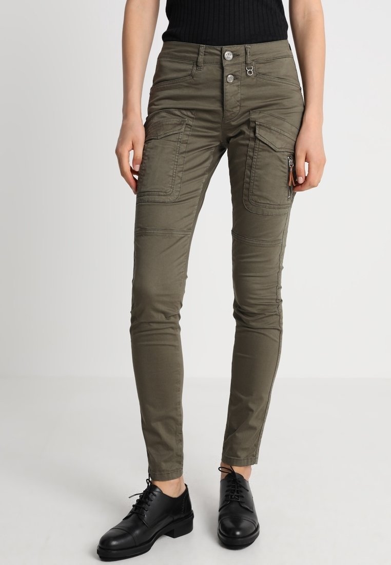 Pantalon cargo slim fit vert olive avec multiples poches, détails zips et boutons, porté avec des chaussures noires à lacets et un top sans manches noir.