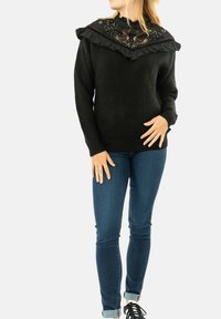 Maglione nero lavorato a maglia con colletto a volant e dettagli ricamati, abbinato a jeans blu e scarpe stringate nere.