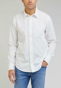 Chemise blanche à col classique en coton, avec des manches longues et un logo subtil ; associée à un jean en denim bleu.