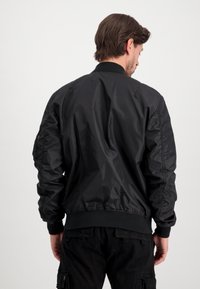 Black bomber jacket with a smooth, shiny finish, ribbed cuffs and hem, and a zip pocket on the left sleeve. Soft texture, tailored fit.