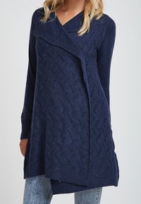 Navy blue knitted tunic with a wave pattern, V-neck, and asymmetrical hem. Long sleeves with ribbed cuffs and textured fabric.