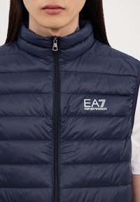 Jūras zilo polsterēto veste ar augstu apkakli, ar rāvējslēdzēju un elastīgām detaļām, ar baltu EA7 Emporio Armani logo uz krūtīm.