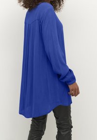 Långärmad blå blus med en strukturerad yta, rundad nederkant och veckade manschetter, kombinerad med slimmade svarta byxor.