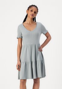 Vestido verde claro canelado com mangas curtas, decote em V e saia em camadas. Apresenta bordas drapeadas e um corpete ajustado.