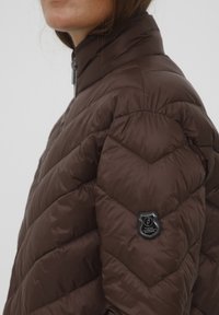 Brūns pūkains mētelis ar chevron rakstu, augstu apkakli un priekšējo rāvējslēdzēju. Uz kreisā piedurkne ir melna logo uzlīme. Gluda tekstūra.