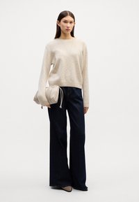 Beige gebreide trui met lange mouwen, gecombineerd met donkerblauwe wijduitlopende jeans. Houdt een lichtbeige clutch met geplooide details vast.