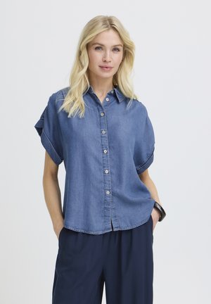 Femme blonde portant une chemise en jean bleue à manches courtes boutonnée et un pantalon bleu marine, debout les mains dans les poches devant un fond uni.