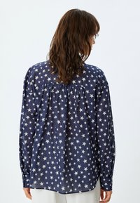 Blusa azul marino con patrón de estrellas blancas, mangas largas, detalle de yugo fruncido en la parte de atrás y puños abotonados. Tejido ligero.