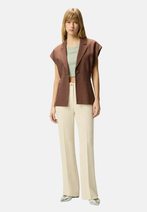 Giovane donna con blazer marrone senza maniche, top corto verde chiaro, pantaloni crema a gamba larga e scarpe metallizzate appuntite, in piedi su sfondo semplice.