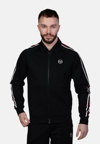 Čierna bunda na zips vyrobená z hladkej látky. Má červené a biele prúžky na rukávoch, rebrované manžety a logo na hrudi.