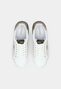 Une paire de baskets blanches Guess avec des accents dorés et des lacets, vue de dessus sur un fond blanc uni.