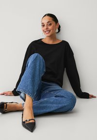 Camisa negra de manga larga combinada con unos vaqueros de denim azul de corte holgado y unos zapatos planos negros de punta. Tejido suave y silueta relajada.