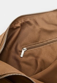 Intérieur d'un sac fourre-tout en tissu brun avec une poche zippée en métal argenté. Texture lisse, fermeture éclair et coutures visibles le long des bords.