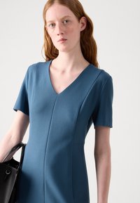Blue short-sleeved dress with a V-neck, featuring a fitted design and seam details. Model holds a black textured handbag.