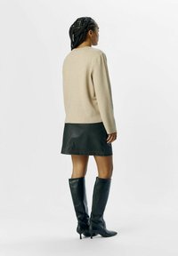 Pull en tricot beige à manches longues, associé à une jupe en simili cuir noire et des bottes noires à talons hauts jusqu'aux genoux. Fond uni.