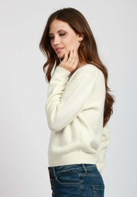 Donna con lunghi capelli castani che indossa un maglione bianco e jeans blu, mentre si tocca delicatamente il viso con la mano destra, su uno sfondo neutro.