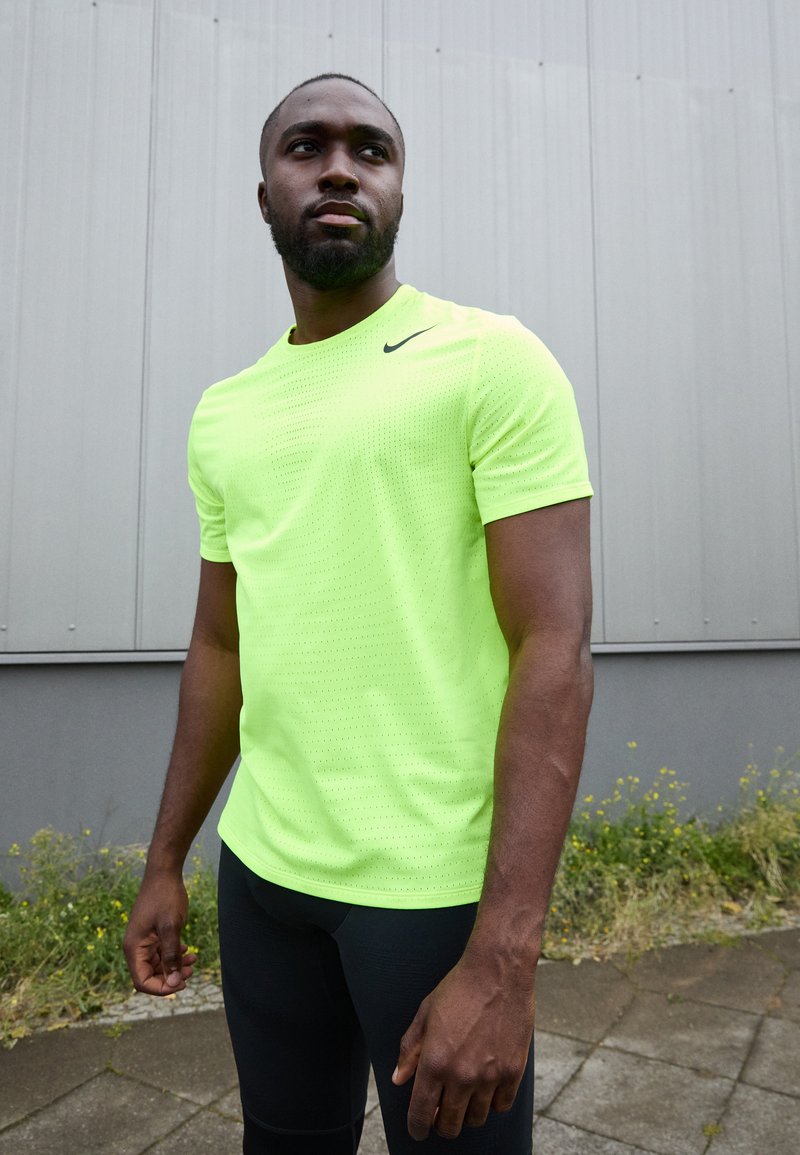 Neonowy żółty T-shirt sportowy z okrągłym dekoltem i krótkimi rękawami, z małym czarnym logo, w połączeniu z czarnymi dopasowanymi legginsami.