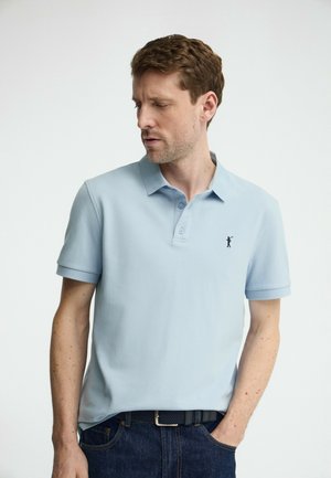 Man draagt een lichtblauw poloshirt met een klein, donker golflogo op de borst, kijkt naar beneden en naar links, met één hand in zijn zak.