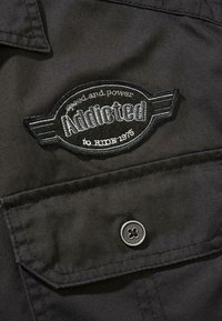 Črna tkanina s kratkimi rokavi, ki vključuje osmišljeno ovalno aplikacijo z napisom "Addicted" v srebrni barvi ter "speed and power to RIDE 1973." Metalni gumbni detajl na žepu.