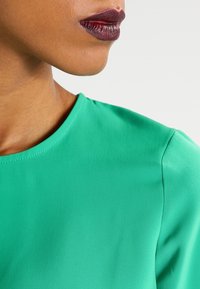 Tessuto verde e liscio con scollatura rotonda, inquadratura ravvicinata della spalla e parte del collo.