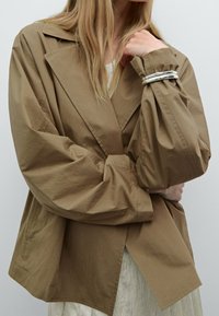 Frau trägt eine oversized braune Jacke mit breiten Revers und silbernen Armreifen am Handgelenk, posiert vor einem schlichten, hellen Hintergrund.