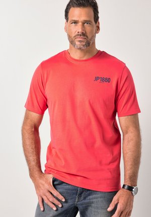 Mężczyzna z krótkimi ciemnymi włosami i brodą, ubrany w czerwoną koszulkę JP1880 i niebieskie dżinsy, stojący na tle jednolitego jasnego tła.