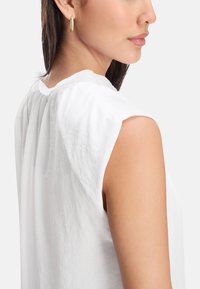 White sleeveless blouse with a pleated back, made of lightweight fabric. Smooth texture and round neckline. Simple, clean design.