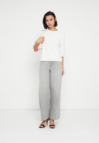 Λευκός πλεκτός πουλόβερ με κοντά μανίκια, συνδυασμένο με γκρι ανοιχτόχρωμο τζιν με φαρδιά μπατζάκια. Το μοντέλο κρατά μια μικρή κρεμ τσάντα και φορά μαύρες γόβες.