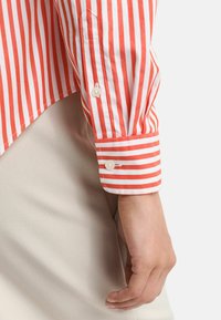 Camicia a righe rosse e bianche con polsini abbottonati, caratterizzata da una texture liscia e una vestibilità sartoriale, abbinata a un tessuto neutro nella parte inferiore.