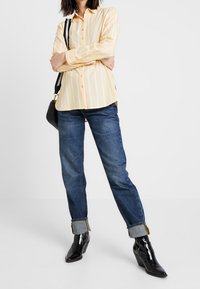 Camisa de manga larga a rayas amarillas y blancas, combinada con unos jeans oscuros de tiro alto y botas de tacón negras a la muñeca, junto con un bolso negro de hombro.