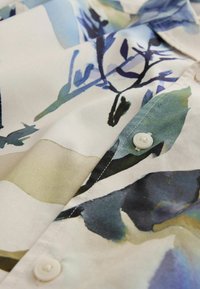 Gros plan sur le tissu d'une chemise boutonnée avec un motif floral abstrait dans des tons de bleu, vert et beige.