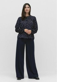 Marineblauwe blouse met polkadots in lichte stof, met een geplooide halslijn en elastische manchetten; gecombineerd met wijde, donkere broeken.