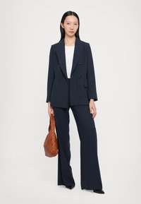 Woman standing wearing navy double-breasted blazer and wide-leg trousers with white top, holding brown woven handbag against white background.
