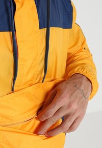 Chaqueta cortavientos amarilla y navy con media cremallera, bolsillo frontal y cordones. Tejido texturizado con puños y dobladillo elásticos.