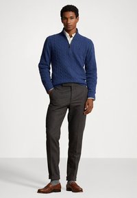 Μπλε πουλόβερ με πλέξη καλτσοδέτας και φ�ερμουάρ στο ένα τέταρτο, φορεμένο πάνω από ένα λευκό πουκάμισο. Το ντύσιμο ολοκληρώνεται με γκρι παντελόνι και καφέ δερμάτινα παπούτσια.