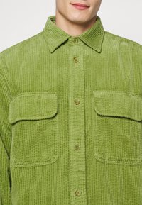 Camicia in corduroy verde con colletto, due tasche sul petto e chiusura con bottoni. Tessuto con texture a righe verticali.