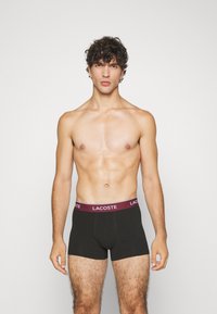 Fekete boxer alsónadrág bordó rugalmas derékrésszel, rajta a "LACOSTE" logóval. A modellen tónusos testalkat van, és mezítelen a felsőteste.