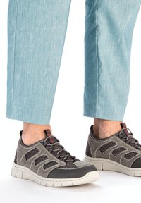 Des chaussures de sport grises avec des panneaux en maille, des accents foncés contrastants et des éléments rouges. Portées avec un pantalon court légèrement texturé bleu clair.