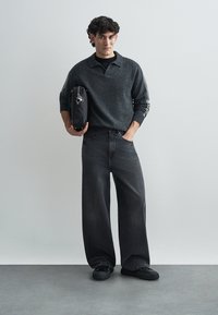 Donkergrijze gebreide trui met een kraag, gecombineerd met zwarte jeans met wijde pijpen. Zwarte tas met metalen details, zwarte platte schoenen op een neutrale achtergrond.