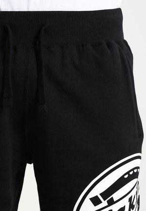 Czarne spodnie dresowe z elastycznym pasem i sznurkiem, z dużym białym okrągłym nadrukiem na górnej części uda.