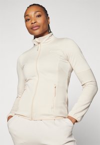 Helle beige Zip-Jacke aus weichem, elastischem Material. Mit gerippten Details an den Ärmeln und Reißverschlusstaschen.