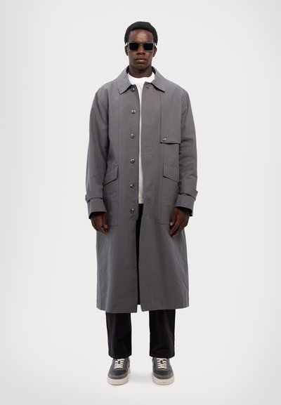 Gray long coat made of cotton, featuring a button-up front, two side pockets, and a relaxed fit. Paired with a white shirt and dark pants.