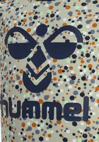 Grüner Stoff mit einem Muster aus einer Mischung kleiner hexagonaler und kreisförmiger Formen in Orange, Blau und Weiß, versehen mit einem hervorgehobenen marineblauen Logo.