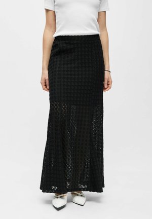 Femme portant une jupe longue noire texturée semi-transparente associée à un haut blanc à manches courtes et des chaussures blanches à bouts pointus avec des nœuds.