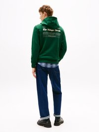 Sweatshirt verde com capuz, com "The Hunger Herald" impresso nas costas, combinado com jeans de ganga azul-escuro com as mangas dobradas e sapatos pretos.
