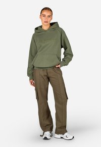 Ελαιόχρωμο φούτερ με μπροστινές τσέπες και κεντημένο λογότυπο, συνδυασμένο με ελαιόχρωμες cargo παντελόνες με πλάγιες τσέπες και λευκά αθλητικά παπούτσια.